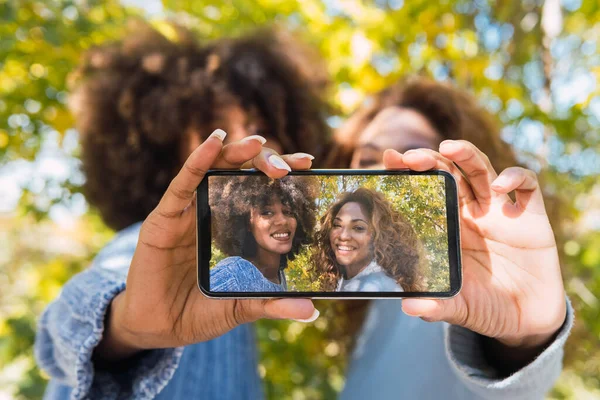 Kıvırcık saçlı, neşeli genç Afro siyah arkadaşlar ön kamerayla akıllı telefonla selfie çekiyorlar. Selfie çekerken gülümseyen iki Afro-Amerikalı kadın.