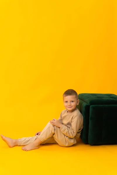 Um menino louro está sentado ao lado de um pufe verde e olhando para a câmera. Fundo amarelo, espaço de cópia — Fotografia de Stock