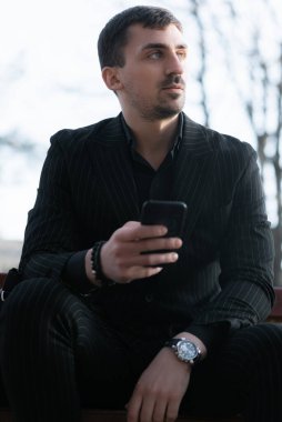 Siyah takım elbiseli bir işadamı bankta oturuyor ve akıllı telefon kullanıyor. İş ve finans kavramı