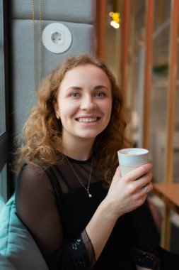 Gülümseyen kızıl saçlı kız elinde bardakla kafede içki tutuyor.