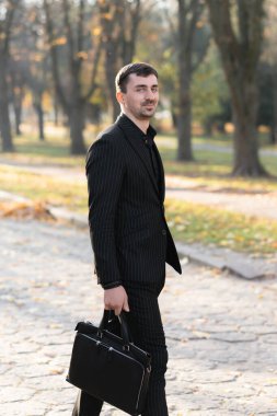 Genç bir işadamı parkta yürüyor, gülümsüyor ve kameraya bakıyor. İşten sonra dinlen.