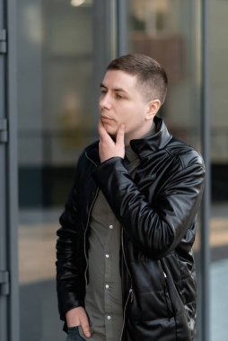 Siyah deri ceketli bir adam yolda yürürken çenesine dokunuyor. Sakallı bir adamın portresi.