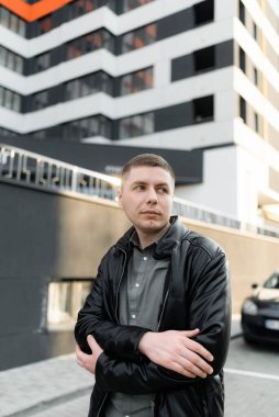 Bir adam otoparkta duruyor, kollarını kavuşturmuş, yan tarafa bakıyor. Sokak portresi