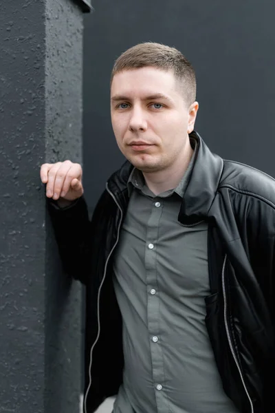 A young man in a black jacket and green shirt is looking at the camera standing near the wall. Street portrait