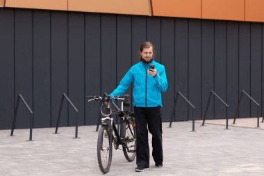 Sosyal medyada gezinen yakışıklı genç bir adam bisikletine yaslandı. Bisikletinin yanında durup gülümseyen ve akıllı telefon kullanan bir adamın portresi.