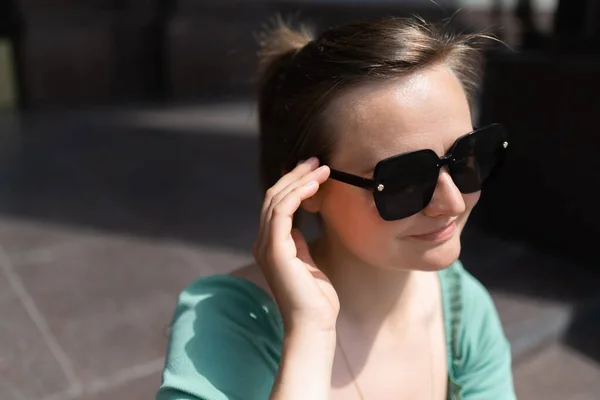 A confident woman wearing a cyan blouse and sunglasses is looking aside. Summer shot on the street background.