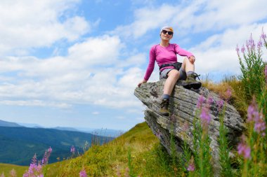A young female hiker sits on a rock in the mountains near chamaenerion angustifolium flowers