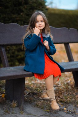 An attractive child is sitting on a park bench wearing a red dress and a blue coat. Fashion portrait