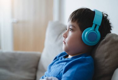Kulaklık takan ve derin düşüncelere dalmış çocuk portresi, müzik dinleyen mavi tişörtlü okul çocuğu, evdeki oturma odasında rahat rahat oturan tatlı çocuk.