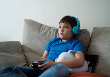 Kulaklık takan ve video oyunu oynayan portre çocuğu. Çocuk evde oyun konsolu tutuyor, genç çocuk kanepede oturuyor ve hafta sonları tek başına eğleniyor.