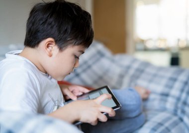 Portre çocuk kanepede oturup tablette çizgi film izliyor, dijital ekranda oyun oynayan sevimli çocuk, kapalı alanda kendi başına rahatlayan çocuk, teknoloji ağlı çocuklar.