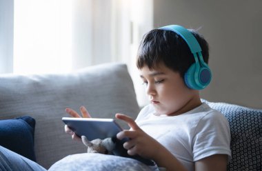 Çocuk kanepede oturmuş tablette çizgi film seyrediyor tatlı çocuk pencerenin yanında oturmuş arkadaşlarıyla oyun oynuyor., 