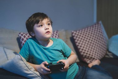 Video oyunu kontrolü elinde tutan mutlu bir çocuğun portresi. Evde online oyun oynayan bir çocuk, kanepede oturan genç bir çocuk bir oyuncakla eğleniyor ve akşam okuldan sonra dinleniyor.