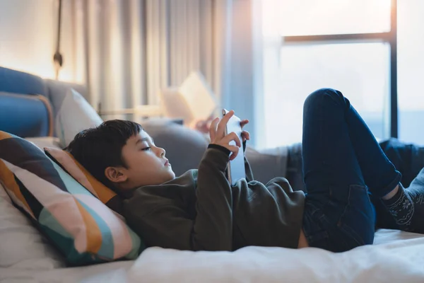 幸せな子供はベッドの中に横たわっているタブレットは漫画を見て デジタルパッド上の友人とチャット かわいい若い男の子はインターネット上でオンラインゲームをプレイし 子供は家族と一緒に家でリラックス — ストック写真