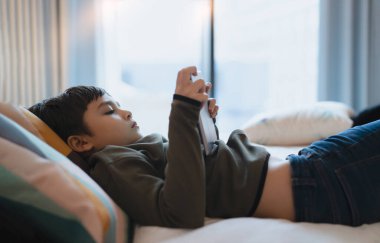 Mutlu Çocuk elinde tabletle yatakta yatıp çizgi film seyrediyor ve dijital ekranda arkadaşlarıyla sohbet ediyor. İnternette oyun oynayan sevimli genç çocuk, ailesiyle evde dinleniyor.