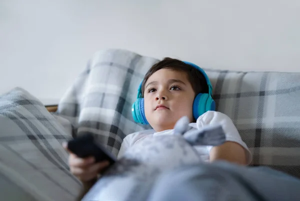 幸せな子供の肖像はヘッドフォンを着用し テレビを見上げ 子供はソファに座ってリモコンを握って 幸せな若い男の子は自宅のリビングルームで音楽を聞いてリラックス — ストック写真