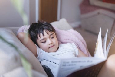 Mutlu okul çocuğu kanepede uzanıp pencereden sabah ışığı parlayan bir kitap okuyor. Çocuk çocuk kitap okuyor hafta sonu evde dinleniyor, olumlu çocuk konsepti