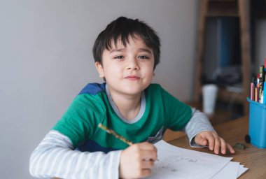 Mutlu çocuk kalem çiziyor ya da kağıda çizim yapıyor, sevimli çocuk kameraya bakıyor, gülümsüyor masaya oturuyor, ödevlerini yapıyor, çocuk evde sanat ve zanaat faaliyetlerini seviyor, eğitim kavramını uyguluyor.