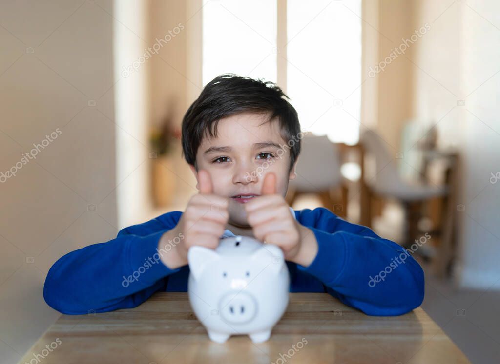 Happy School boy putting thumbs up on piggy bank and looking at camera with smiling face.Child showing money saving box.kid Learning financial responsibility and planning about saving money for future