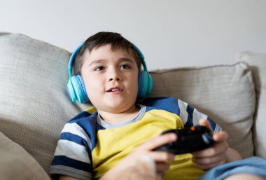 Kulaklık takan ve video oyunu oynayan portre çocuğu. Çocuk evde oyun konsolu tutuyor, genç çocuk kanepede oturuyor ve hafta sonları tek başına eğleniyor.