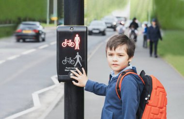 School kid pressing a button at traffic lights on pedestrian crossing on way to school. Child boy with backpack using traffic signal controlled pedestrian facilities for crossing road. clipart