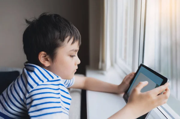 映画のポートレート若い男の子がタブレット上でゲームをプレイします ソファの上に座っている光が窓から輝いて 子供はインターネット上でゲームをプレイ 子供は家で宿題をオンラインで行う — ストック写真