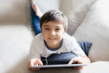 Yüksek anahtarlı portre genç çocuk tablette oyun oynuyor kanepede oturuyor pencereden yansıyan ışıkla, çocuk internette oyun oynuyor, evde ödev yapan sinematik çocuk