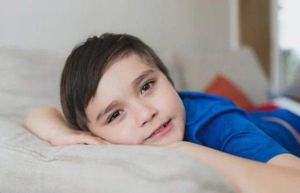 笑顔のカメラを見上げソファに寝そべっている幸せな男の子 顔を閉じて晴れた日の春や夏に窓から光が輝くリビングルームで休んでいる子供 週末に家でリラックスする子供 — ストック写真