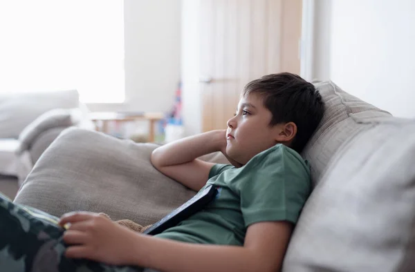 Kid with remote control and looking up with curious face,Young boy sitting on sofa watching cartoon on TV, Portrait Child lying down on couch relaxing in living room after back from school.