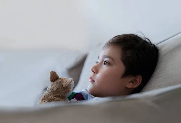 Close up face kid sitting on sofa with dog toy watching TV, Young boy   sitting on couch looking out deep in thought. Positive Child relaxing at home on weekend