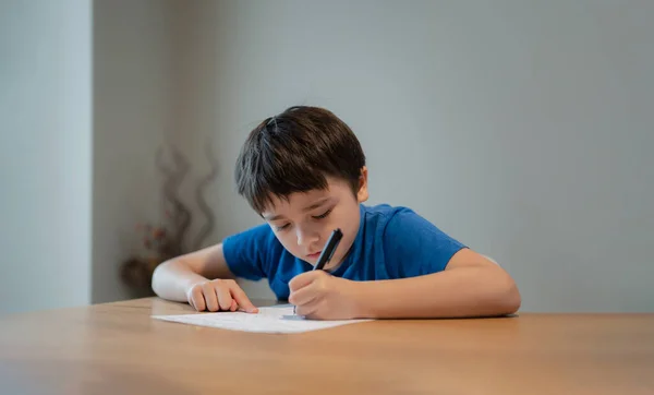 Kid siting on table doing homework,Child boy holding black pen writing on white paper,Young boy practicing English words at home. Elementary school and home schooling, Distance Education concept