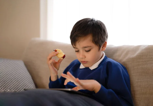Happy schoolboy eating red apple while playing game online with friend on tablet,Kid using internet sending homework to the teacher,Positive child sitting on sofa relaxing alone in living room in the morning