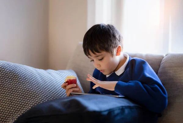 Happy schoolboy eating red apple while playing game online with friend on tablet,Kid using internet sending homework to the teacher,Positive child sitting on sofa relaxing alone in living room