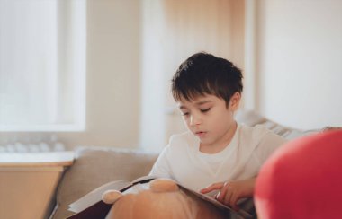 Happy School kid sitting on sofa reading a book with morning light shining from window. Child boy reading story relaxing at home on weekend, Positive children concept