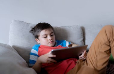 School kid using tablet readying story from internet, Happy boy sitting on sofa doing studying online learning  at home, Child holding digital pad using wifi playing game or chatting with friends.
