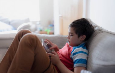 School kid using tablet readying story from internet, Happy boy sitting on sofa doing studying online learning  at home, Child holding digital pad using wifi playing game or chatting with friends.