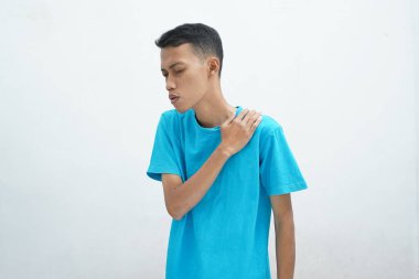 Asyalı genç adam mavi tişört giyiyor. Beyaz arka planda denemekten omuz ağrısı çekiyor.