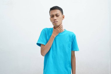 Mavi tişört giyen genç Asyalı adamın boğazı ağrıyor ve beyaz arka planda izole edilmiş boynuna dokunuyor. Yutması zor.
