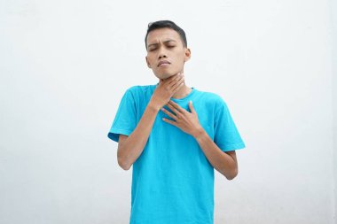 Mavi tişört giyen genç Asyalı adamın boğazı ağrıyor ve beyaz arka planda izole edilmiş boynuna dokunuyor. Yutması zor.