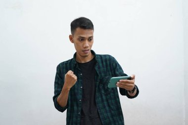 Çekici genç Asyalı adam koyu yeşil günlük kıyafetler giyiyor telefonunda iyi haberler var, mutlu gülüyor, gülüyor, beyaz arka planda muzaffer bir ifade var.