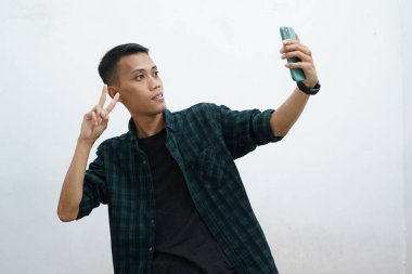 Koyu yeşil giysili genç Asyalı adam beyaz arka planda görüntülü görüşme yapıyor ve selfie çekiyor.