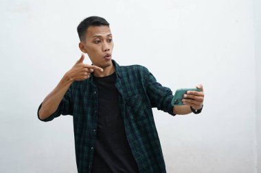 Çekici genç Asyalı adam koyu yeşil günlük kıyafetler giyiyor telefonunda iyi haberler var, mutlu gülüyor, gülüyor, beyaz arka planda muzaffer bir ifade var.