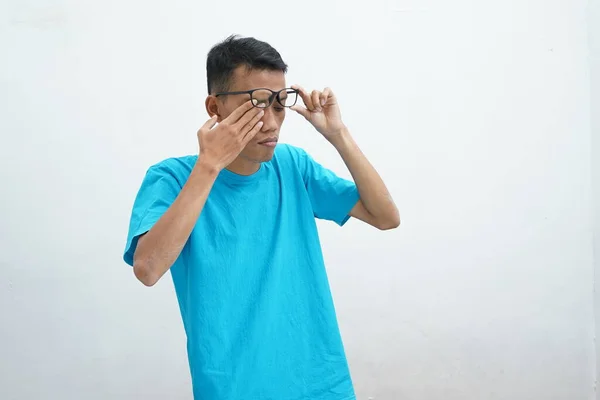年轻的亚洲人穿着不健康的蓝色T恤 戴着眼镜 揉着眼睛 视力模糊 眼睛干瘪 被白色背景隔离 健康问题概念 — 图库照片