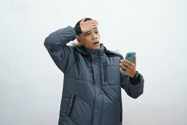 İzole edilmiş beyaz arka planda kışlık ceket giyen Asyalı bir adam. Elleri başının üstünde stresli bir akıllı telefon kullanmak.