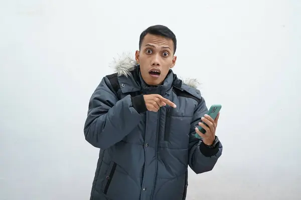 亚洲人穿着冬季夹克 背景是孤立的白色 一边喊着一边看着相机 一边指着他的智能手机 这都是坏消息 — 图库照片
