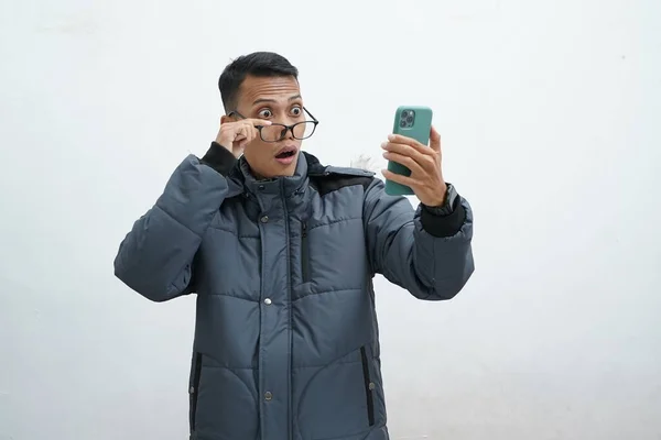 亚洲人穿着冬季夹克 戴着眼镜 罩在孤立的白色背景上 他从智能手机里得到的好消息使他看起来很惊讶 — 图库照片