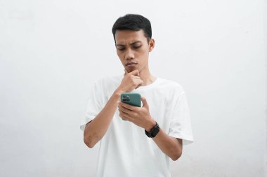 Beyaz tişörtlü Asyalı adam, akıllı telefon ekranına bakarken bir şey düşünüyor, sanki şüpheli bir şey var gibi..