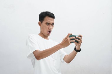 Öfkeli Asyalı adam beyaz tişört giyiyor. Beyaz arka planda Smartphone Tableti 'nde oyun oynuyor..