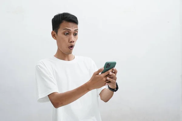 白いTシャツを着たアジア系の男は スマートフォンから届いた良い知らせに驚く 焦点を当てた目でショックを受けたジェスチャーを示す男はスマートフォンを見て — ストック写真