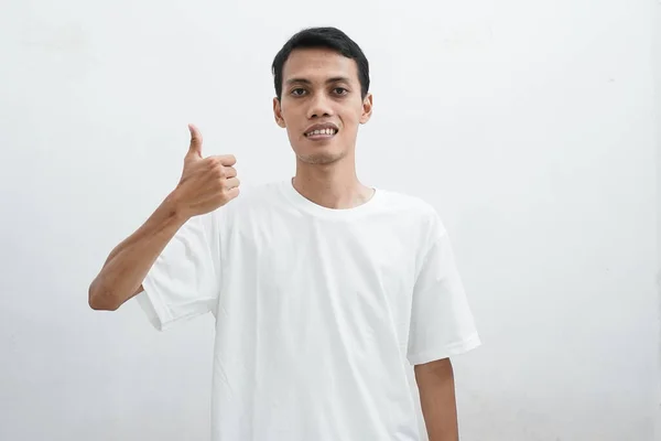 身穿白色T恤的亚洲人一边对着相机微笑 一边竖起大拇指 有选择性地聚焦于白色背景的半身像 — 图库照片